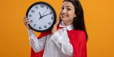 smiling-young-caucasian-superhero-girl-holding-clock-looking-camera-isolated-orange-background-with-copy-space_141793-77239